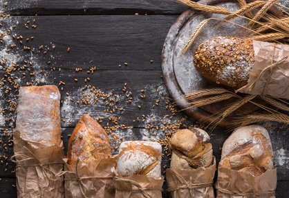 Bread background. Brown and white whole grain loaves wrapped in kraft paper composition on rustic dark wood with wheat ears scattered around. Baking and home bread making concept. Soft toning